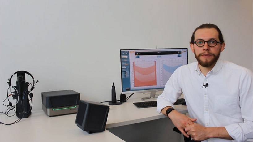 Jack Bennett sat at white desk with the following, and carefully positioned, equipment: 1) Real-ear measurement headset resting on a desktop cradle. 2) Affinity Compact. 3) Loudspeaker placed away from reflective surfaces. 4) Video otoscope standing on its cradle. 5) Computer screen with hearing aid fitting software. 6) Ambient Noise microphone attached to the top of the computer screen.