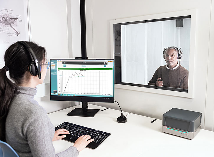 Clinician operating PC keyboard and ACT test screen in the Affinity Suite. The clinician has an Affinity Compact on their desk, and the clinician is looking through a glass window to a client sat on the other side in a booth. The client is wearing headphones and pushing a patient response button.