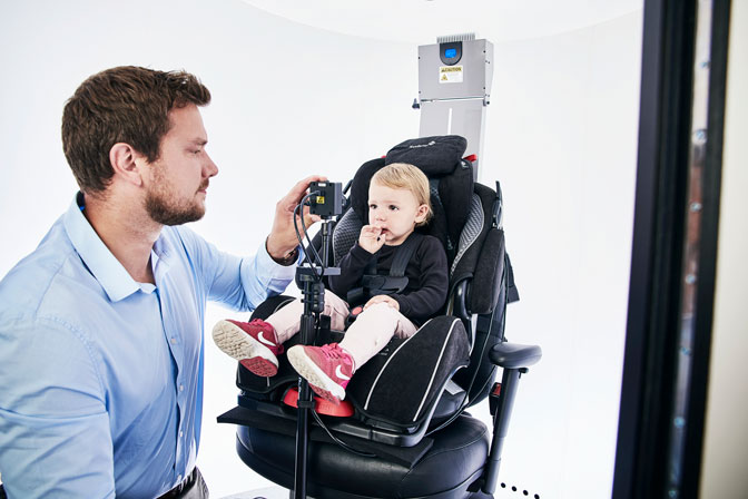 Little girl sitting in car seat, looking toward the in-booth camera. Male clinician operating the in-booth camera.