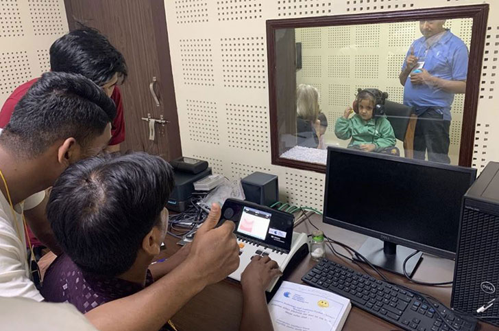 Three Nepali CEHWs sat by audiometer on one side of a window looking into an audiometric booth. A small girl on the other side of the window wearing headphones and accompanied by a Nepali CEHW and a GFCHL representative.