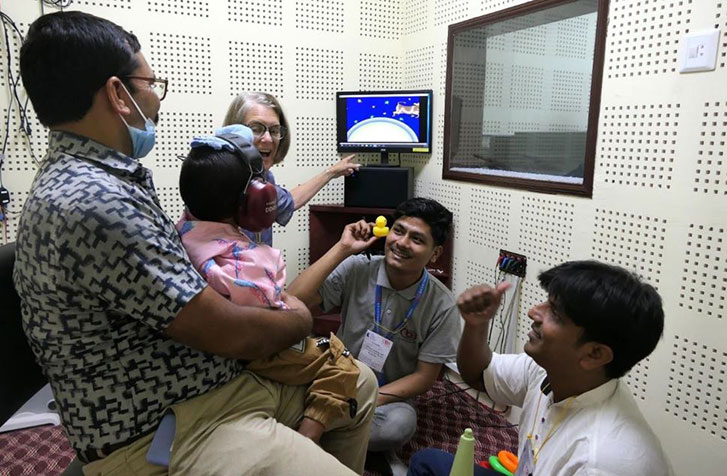 A GFCHL representative and two Nepali CEHWs performing visual reinforcement audiometry in a young child inside an audiometric booth. The child is sat on their father’s lap.