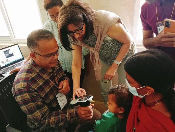 A GFCHL representative is instructing a Nepali CEHW on how to operate the tympanometer which he is holding in his right hand. Two other CEHWs are also present, as is a young boy, who is sat on his mother’s lap, facing toward the CEHW holding the tympanometer.