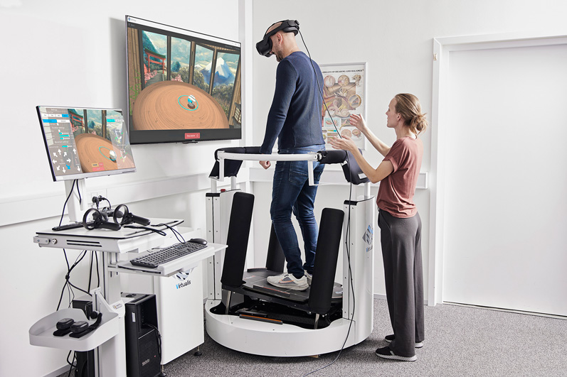 A patient is standing on the enclosed dynamic force plate, which is tilting backward to the right. A clinician is standing behind the patient on the floor at a short distance, with their right hand held up to be ready to support the patient should they fall.