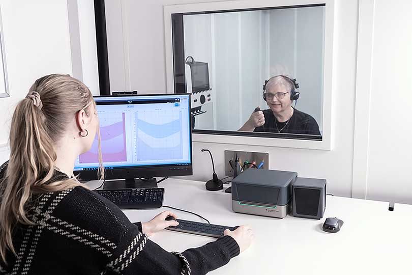 Eine Audiometristin sitzt an einem weißen Schreibtisch mit Audiometer-Tastatur, Affinity Compact, Maus, Lautsprecher, Mikrofon, PC-Tastatur und einem Computerbildschirm, auf dem Audiometrie-Software angezeigt wird. Mit ihren beiden Händen bedient die Audiometristin beide Regler der Audiometer-Tastatur. Sie schaut durch ein Fenster, hinter dem eine Patientin in einer Kabine sitzt. Die Patientin trägt Kopfhörer und drückt eine Patienten-Antworttaste.