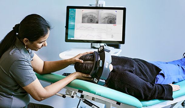 Clinician performing positioning maneuver on patient that is wearing VNG goggles and in a supine position on an examination bed. Computer screen in background showing live eye video and eye movement data