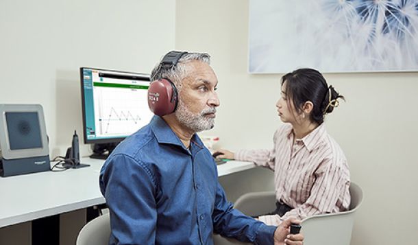 Clinician performing ACT test in client. The client is wearing a blue shirt, has headphones on, and is holding a patient response button. In the background, the clinician is operating the ACT software on a computer screen.