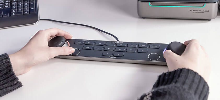 Audiometer-Tastatur auf einem weißen Schreibtisch vor einer PC-Tastatur und dem Affinity Compact. Eine Person bedient mit ihren beiden Händen die beiden Regler links und rechts auf der Tastatur. Zwischen den Multifunktionsreglern befinden sich mehrere Tasten, darunter Ein- und Ausgang für Kanal eins und zwei, Speichern, zwei Tasten zum Wechseln der Frequenzen sowie neun Funktionstasten mit den Bezeichnungen F1 bis F9.