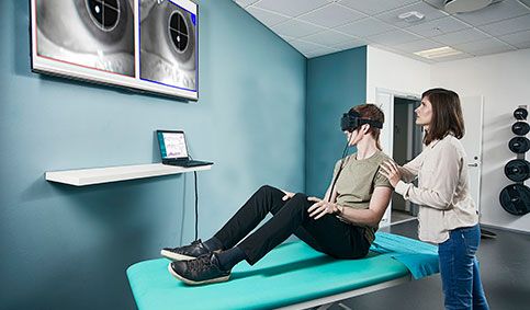 Female clinician performing Advanced Dix-Hallpike maneuver in a young man, who is sat on an examination bed situated in a physical therapy clinic. The man is wearing Video Frenzel goggles with a head sensor mounted. The man is looking toward a computer screen displaying Advanced Dix-Hallpike software. The clinician is looking up toward a large TV screen showing a video image of the patient's eyes.