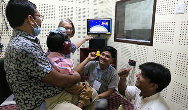 A GFCHL representative and two Nepali CEHWs performing visual reinforcement audiometry in a young child inside an audiometric booth. The child is sat on their father’s lap.