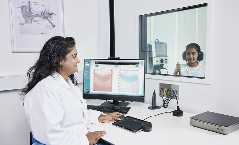 Akustiker führt mit dem Equinox Evo und der Touch Keyboard eine Audiometrie bei einem jungen Mädchen durch, das hinter Glas in einer Audiometriekabine sitzt.