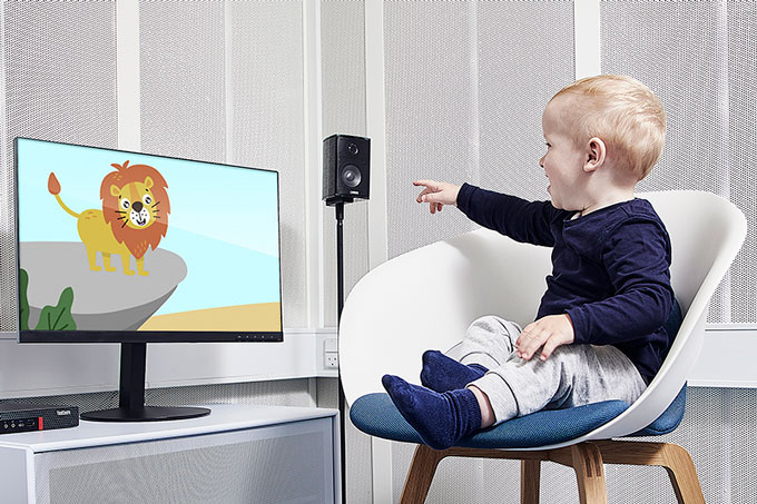 Young boy sat in a white chair on a blue cushion inside a booth. He’s looking and pointing toward a computer screen to his right that is showing a smiling animated lion. The VRA Screen computer and a speaker are located on each side of the computer screen.