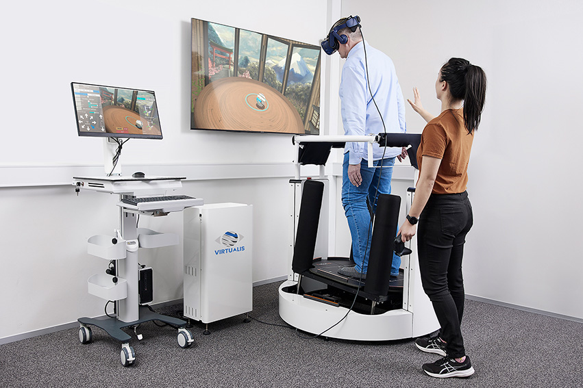A patient is standing on the encLOSed dynamic force plate, which is tilting backward to the right. A clinician is standing behind the patient on the floor at a short distance, with their right hand held up to be ready to support the patient should they fall.