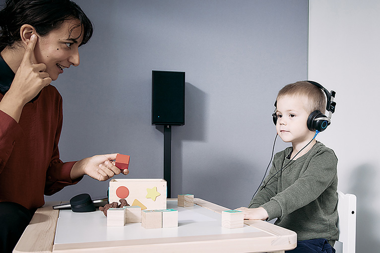 Clinician sat at small table with young child and performing play audiometry. The child is wearing headphones to hear the stimulus. There are Ling-6 blocks and shape puzzles on the table. The clinician has screened the Touch Keyboard from view behind some of the toys on the table.