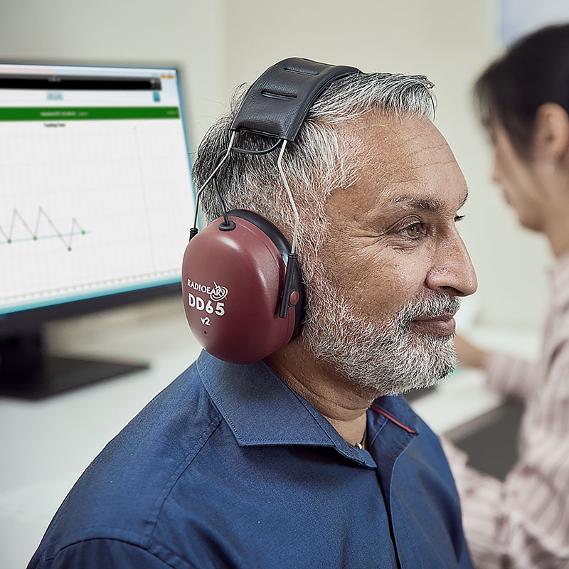 Client wearing Radioear DD65 v2 headphones with clinician operating ACT test software in the background