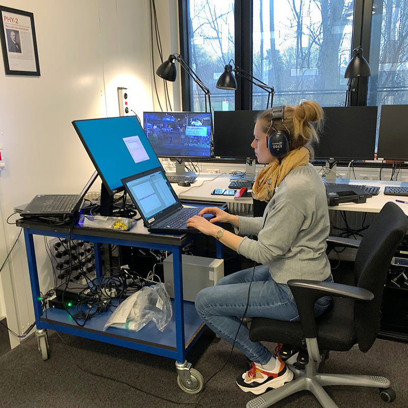 Lisbeth is sat on a chair with wheels and wearing DD65 headphones. Her workstation is full of computer equipment, gadgets, wires, cables and all you could expect for a busy researcher. Lisbeth is working on a laptop, with windows and a view outside to nearby trees to the right of her.