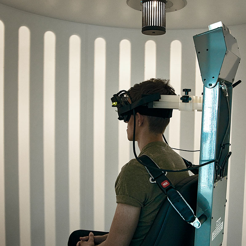 Patient strapped into the enclosed Orion Comprehensive chair and wearing VNG goggles. The light from the optokinetic drum above the patient is displaying a visual pattern across the wall of the booth enclosure.