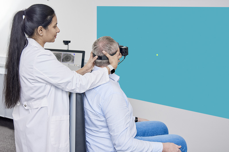 Clinician performing Gaze test in a patient. The patient is sat in the Orion Reclining rotary chair and fixated with VisualEyes VNG goggles, looking toward a dot projected onto a wall. Their body is facing 45 degrees to the right of the stimulus, and the clinician is turning the patients head 45 degrees to the left to look at the stimulus. The positioning is the same for the Smooth Pursuit Neck Torsion test described further down in this article.
