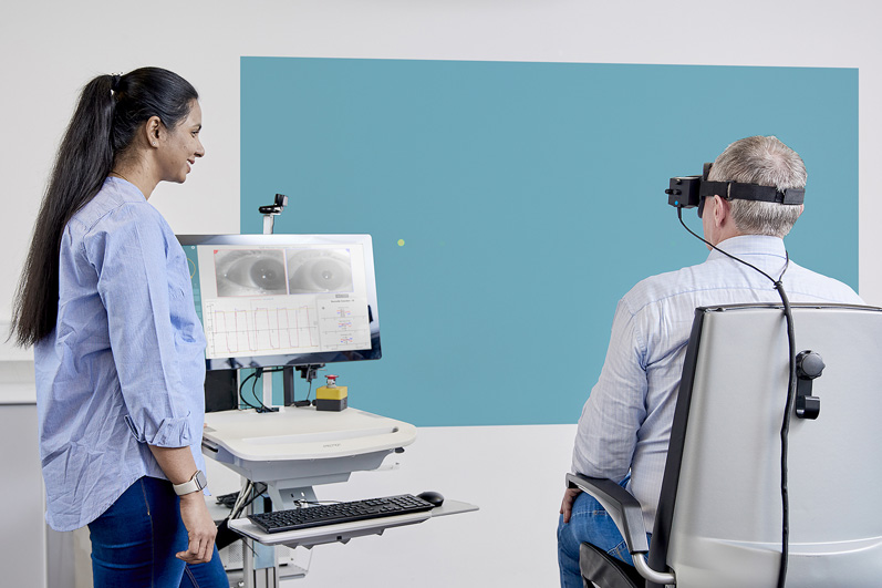 Clinician observing a patient performing self-paced saccades. The patient is sat in the Orion Reclining rotary chair and fixated with VisualEyes VNG goggles, looking toward a dot projected onto a wall.