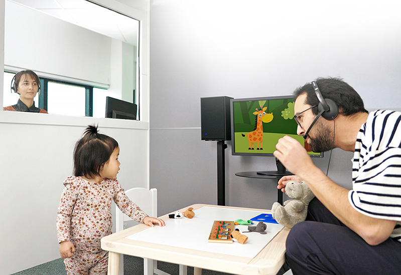 The audiologist controlling the audiometer and the VRA controls is sat outside the audiometric booth, looking through a glass window to see the child and second tester sat inside the audiometric booth. The second tester is playing with the child with a selection of toys. The child is looking to their left at a visual reinforcer displayed on a computer screen, with a speaker next to it.
