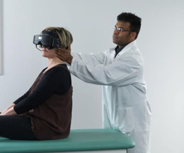 Clinician stood behind patient sat on a green examination bed. The clinician is turning the patient’s head toward the left at a 45-degree angle.