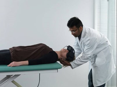 Clinician stood behind patient lying on their back on a green examination bed. The clinician is turning the patient’s head toward the left at a 45-degree angle.
