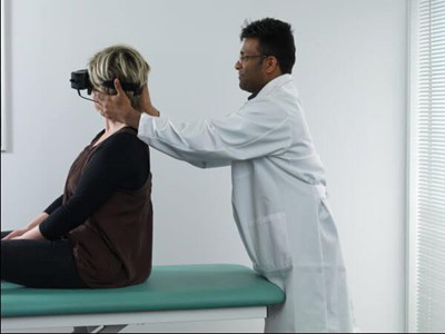 Clinician stood behind patient sat on a green examination bed. The clinician is turning the patient’s head toward the right at a 45-degree angle.