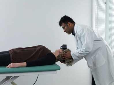 Clinician stood behind patient lying on their back on a green examination bed. The clinician is turning the patient’s head toward the right at a 45-degree angle.