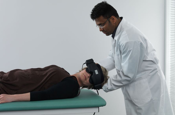 Male clinician performing head roll to the left on female patient. The patient is lying in a supine position and wearing VNG goggles.