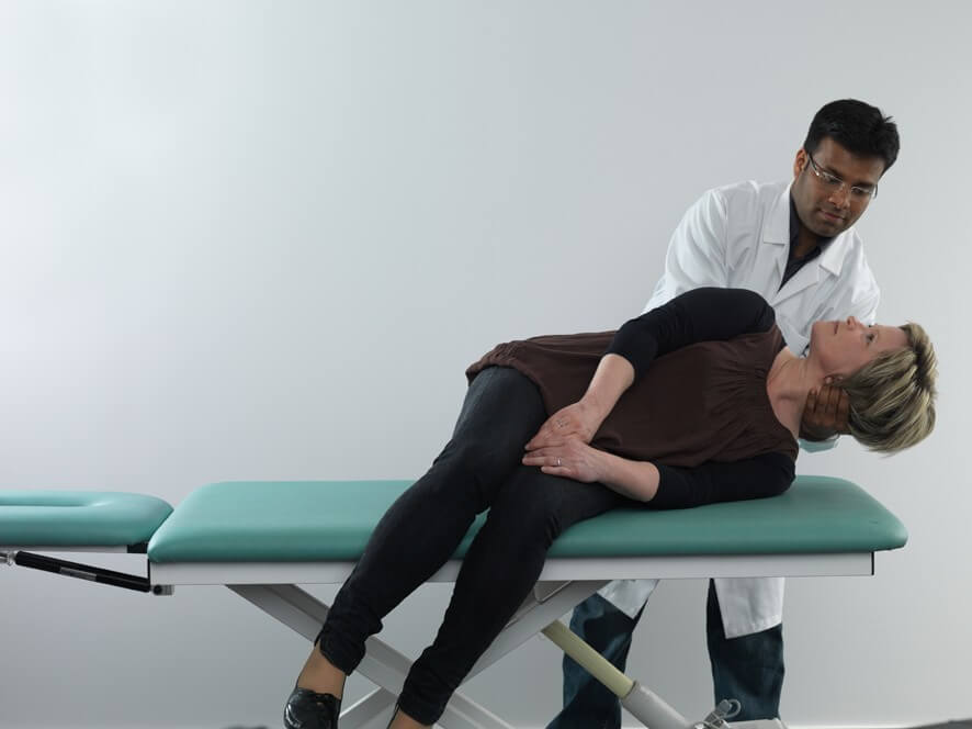 Female patient in black clothes lying on her left side on examination table with legs dangling off the edge of the table. Male clinician stood behind the patient, and is turning her head about 90 degrees toward the right.