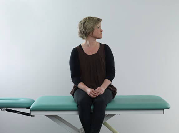 Woman in black clothes sitting on examination table with legs dangling off the edge of the table. She has her head turned about 90 degrees toward the left.