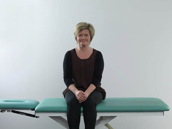 Woman in black clothes sitting on examination table with legs dangling off the edge of the table.