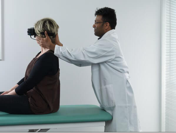 Female patient sitting on examination table with back toward a male clinician. The clinician is turning the patient's head 45 degrees toward the right.