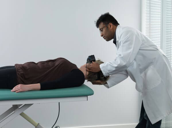 Female patient lying on her back on examination table, with head hanging over the edge of the table. Male clinician is turning the patient's head toward the right.