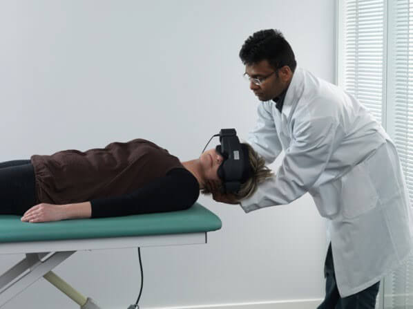 Female patient lying on her back on examination table, with head hanging over the edge of the table. Male clinician is turning the patient's head toward the left.