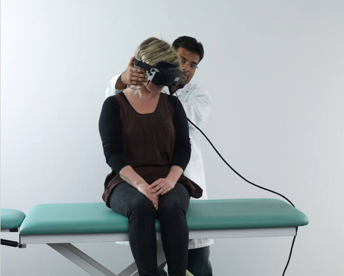 Female patient sitting on examination table with back toward a male clinician, and legs dangling off the edge of the table. The clinician is turning the patient's head 45 degrees toward a downward, left position.