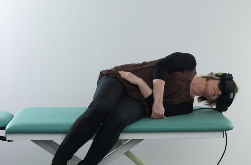 Woman in black clothes, wearing VNG goggles, lying on her left side on green examination table with her legs dangling off the edge of the table.