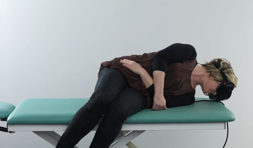 Woman in black clothes, wearing VNG goggles, lying on her left side on green examination table. Her face is pointed down toward the table and her legs are dangling off the edge of the table.