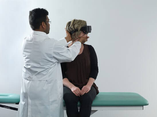 Female patient sat upright on examination table with her legs dangling off the edge of the table. Male clinician stood in front of her, turning her head 45 degrees toward the left.