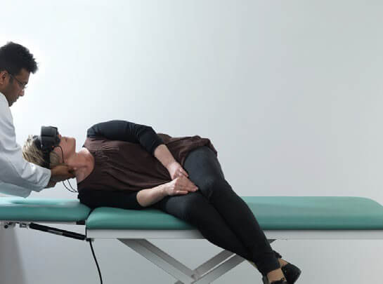 Female patient lying on her right side on examination table with her legs dangling off the edge of the table. Male clinician stood beside her, turning her head 45 degrees toward the left.