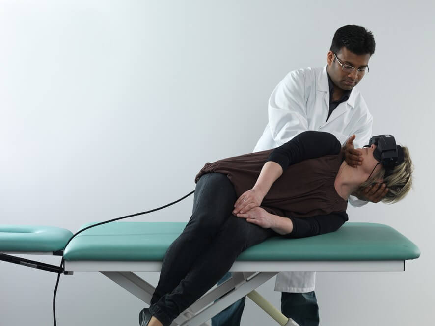 Female patient in black clothes lying on her left on examination table with her legs dangling off the edge of the table. Male clinician stood behind is turning her head 45 degrees toward the right.
