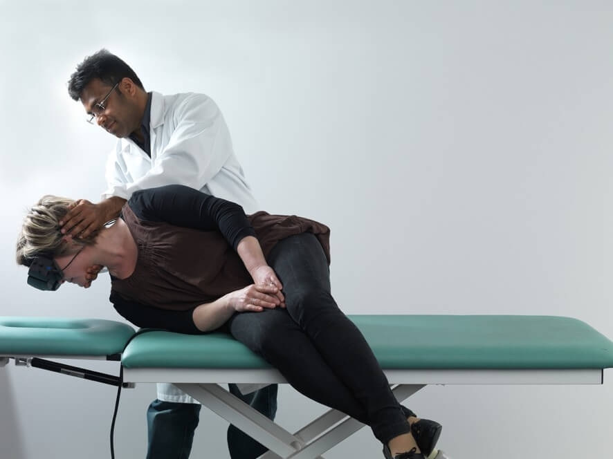 Female patient in black clothes lying on her right on examination table with her legs dangling off the edge of the table. Male clinician stood behind is turning her head 45 degrees toward the right.