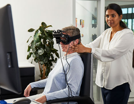Female clinician stood behind female patient, with hands placed on the patient's jaw. The patient is wearing VNG goggles.