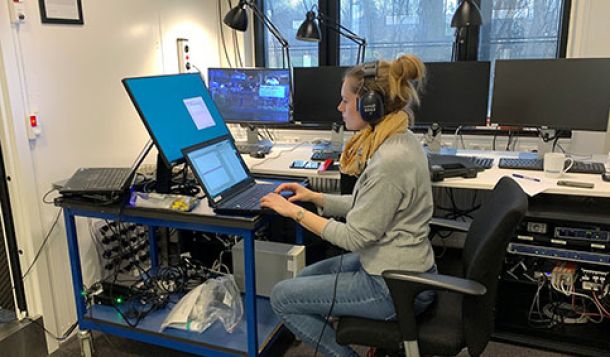 Lisbeth Birkelund Simonsen sat on a chair with wheels and wearing DD65 headphones. Her workstation is full of computer equipment, gadgets, wires, cables and all you could expect for a busy researcher. Lisbeth is working on a laptop, with windows and a view outside to nearby trees to the right of her.