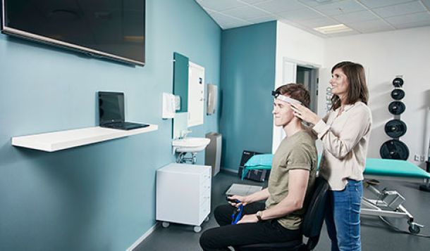 Female clinician in beige shirt stood behind male patient wearing a headband with a sensor mounted. Examination bed and gym equipment visible in the background. The patient is holding a remote and looking toward a laptop screen, performing the gaze stabilization test.