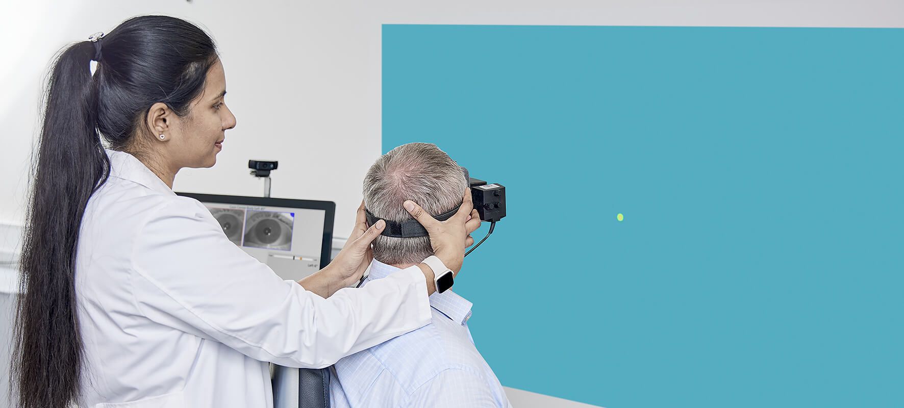 Rear view of patient wearing VNG goggles. A clinician stood behind the patient is turning the patient's head 45 degrees to the left, with the patient looking toward a dot stimulus projected on to the wall in front of them