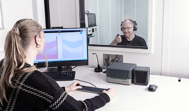 Female clinician sat at a white desk with the Audiometer Keyboard, Affinity Compact, mouse, speaker, microphone, PC keyboard, and a computer screen displaying audiometry software. The clinician is operating the Audiometer Keyboard, with her hands placed on its two dials. The clinician is looking through a window behind which a female patient is sat in a booth. The patient has headphones on and is pressing a patient response button.