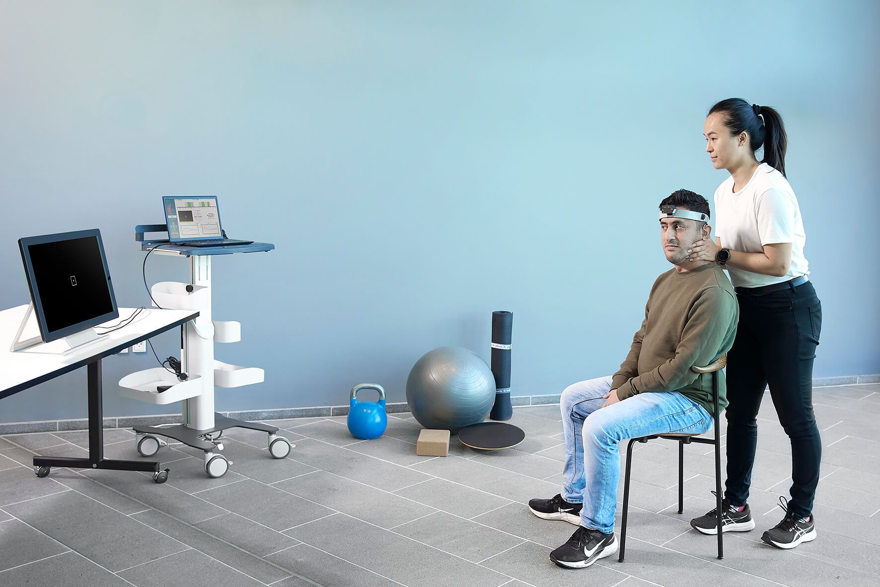 Frontal view of patient wearing VNG goggles. A clinician stood behind the patient is turning the patient's head 45 degrees to the left