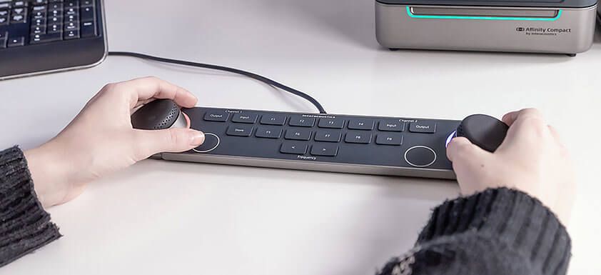 Audiometer Keyboard on a white desk in front of a PC keyboard and the Affinity Compact. A person is operating the keyboard, with their hands placed on the two dials located on the left and right sides of the keyboard. There are several keys between the multi-functional dials, which include and are not limited to input and output for channels one and two, store, two keys to decrease or increase the intensity, and nine F-keys labeled F1 to F9.