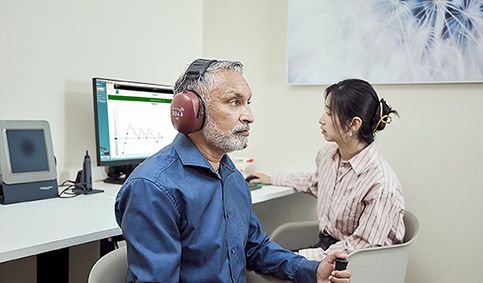 Clinician performing ACT test in client. The client is wearing a blue shirt, has headphones on, and is holding a patient response button. In the background, the clinician is operating the ACT software on a computer screen.