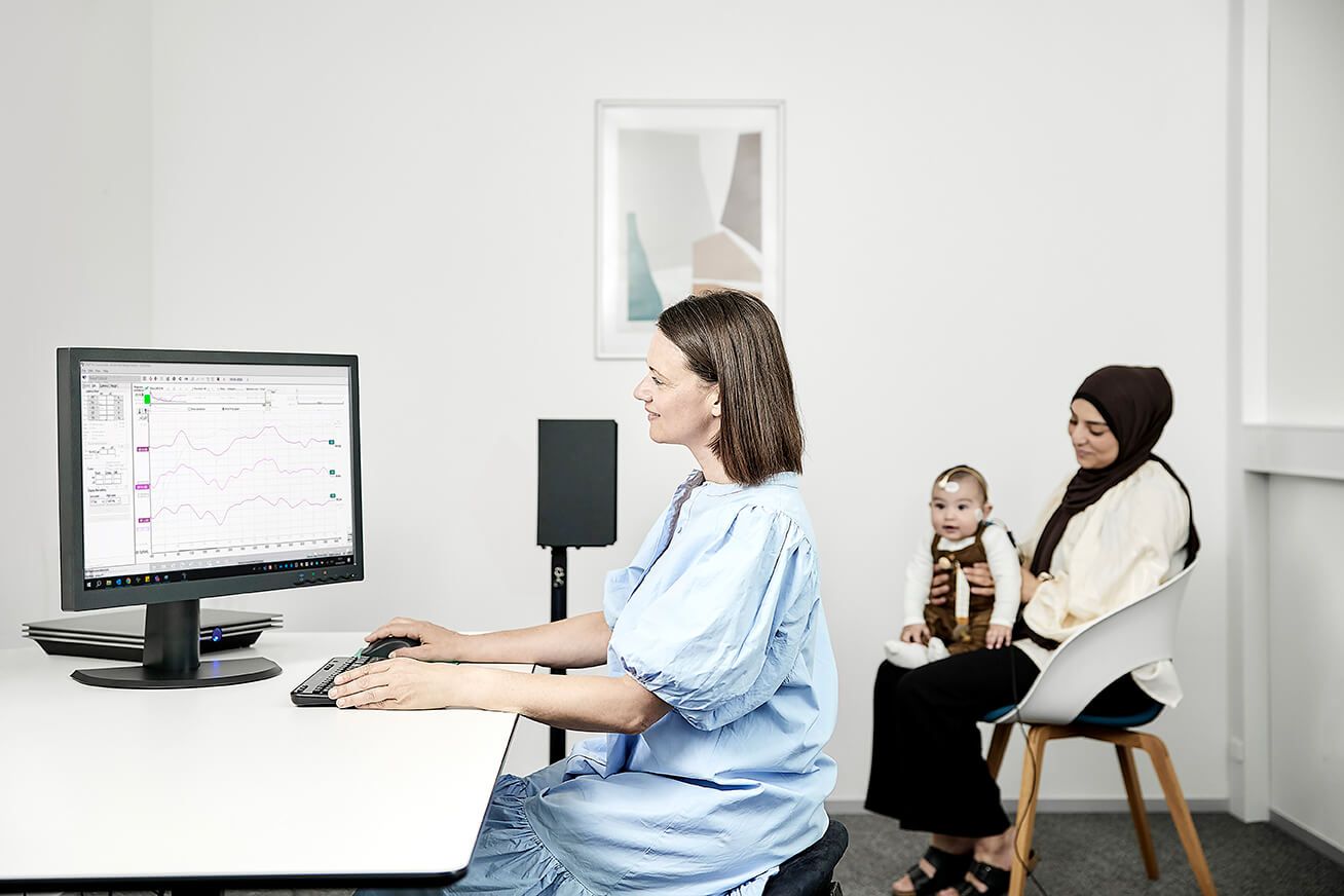 Clinician performing aided cortical testing in a young infant. The clinician is operating the Aided Cortical module on a computer screen, behind which an Interacoustics Eclipse is resting on the desk. In the background, there is a free field setup with one speaker. About a meter and a half away from the speaker, a parent is sitting in a chair with their infant child on their lap. The infant has electrodes placed on their high forehead and cheeks.